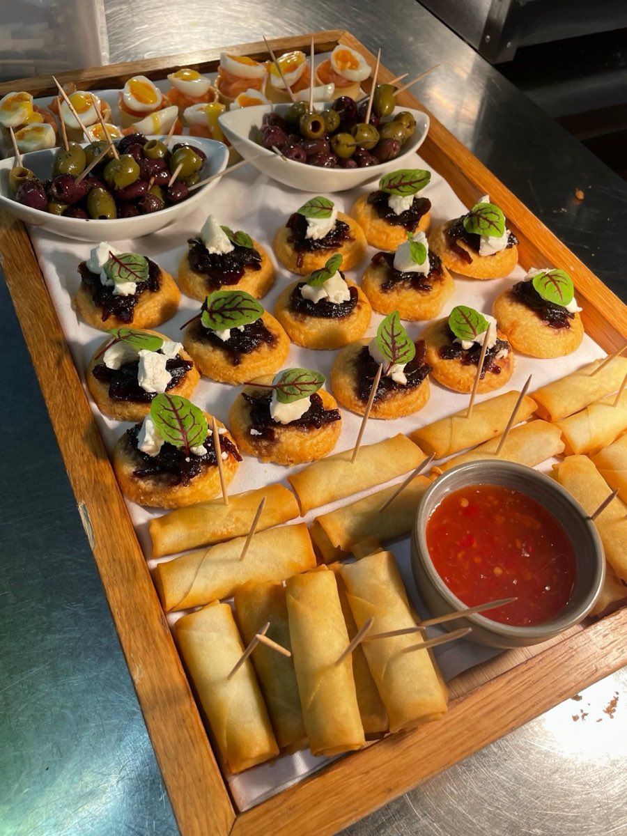 A wooden canapé board of goat's cheese tartlets, spring rolls with chilli dip and marinated olives.
